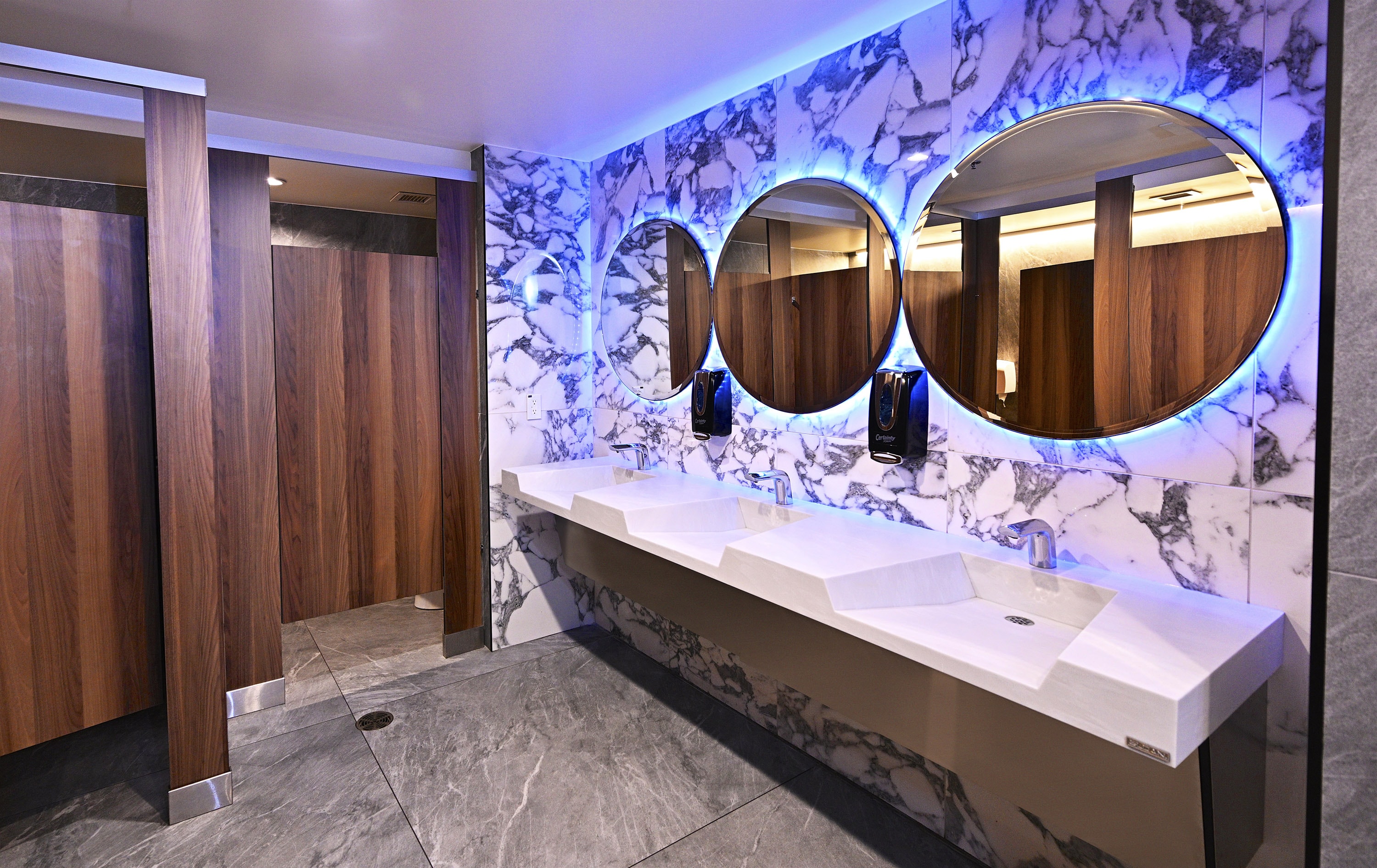 NBA courtside restaurant luxury restroom — marble walls, blue LED backlit circular mirrors, wood stall partitions, and integrated sink countertop